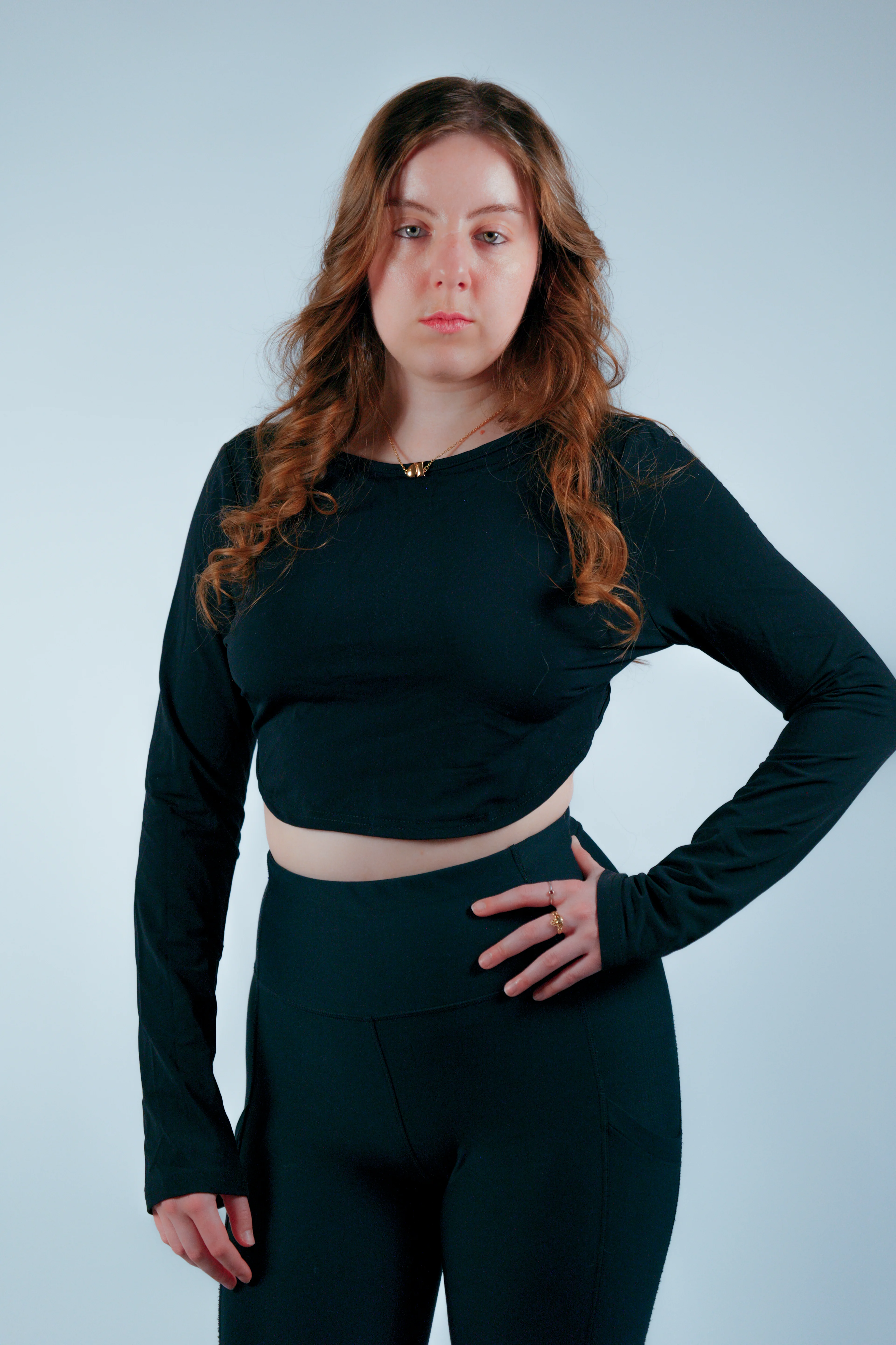 Model in black crop top with gold necklace and pants, hand-on-hip pose on light blue backdrop