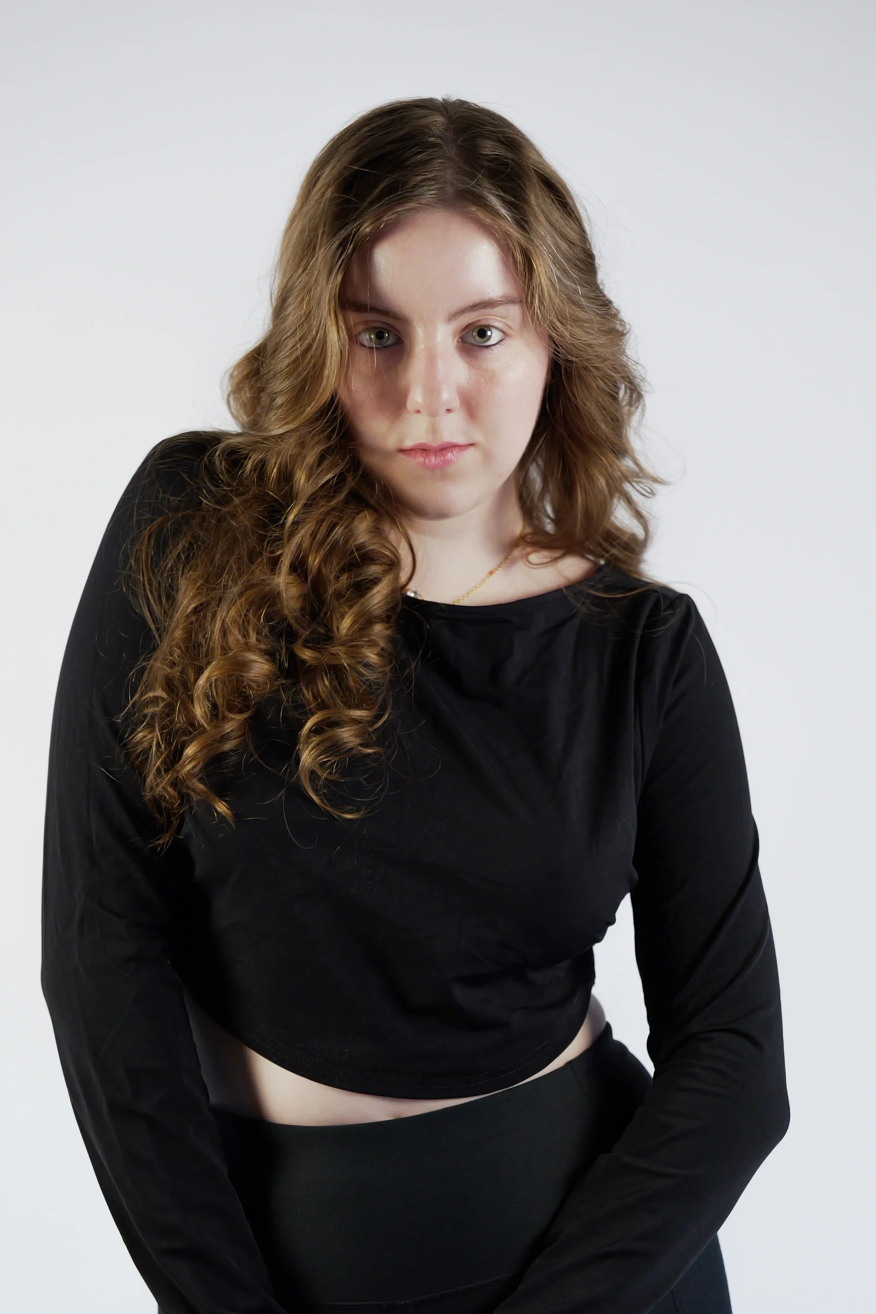 Model in black long-sleeve crop top and pants, seated pose with wavy brown hair on white studio background
