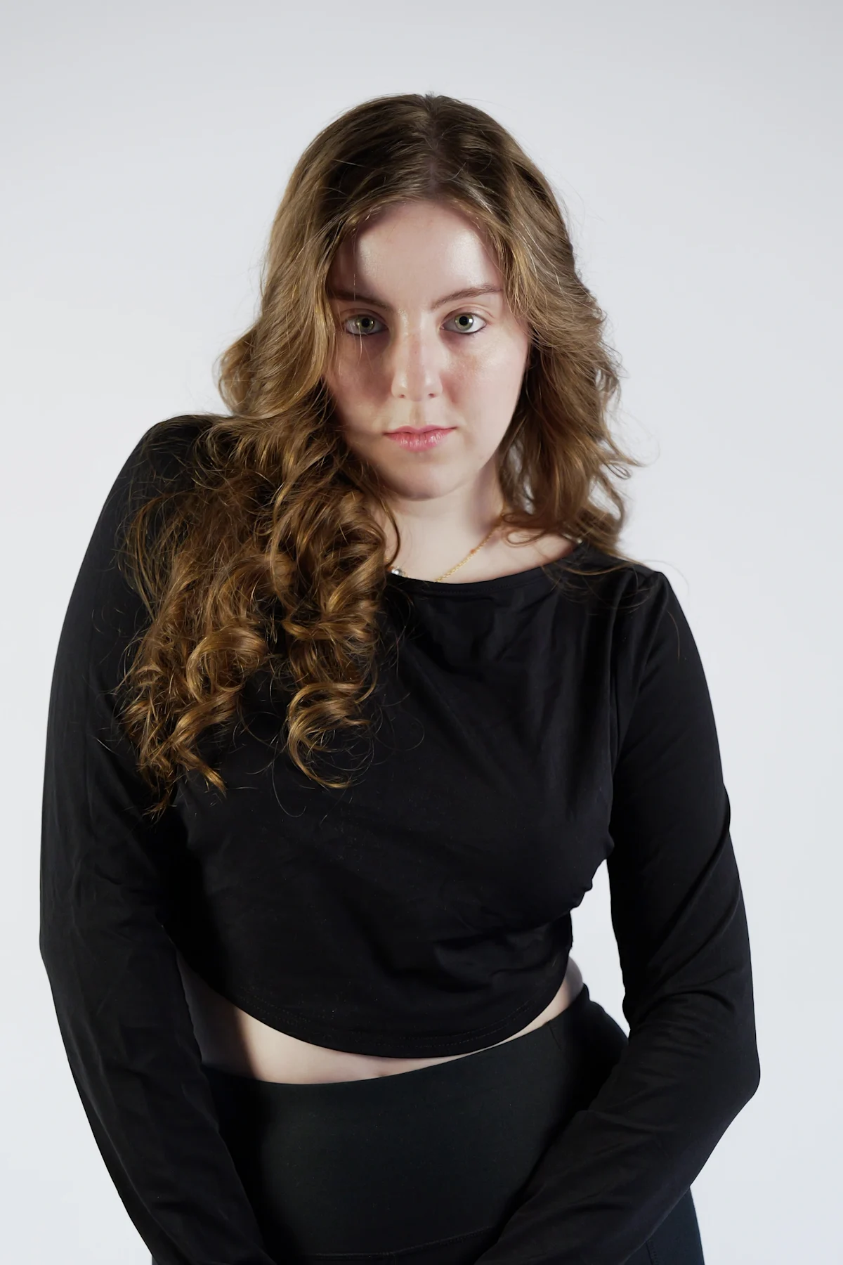 Model in black long-sleeve crop top and pants, seated pose with wavy brown hair on white studio background