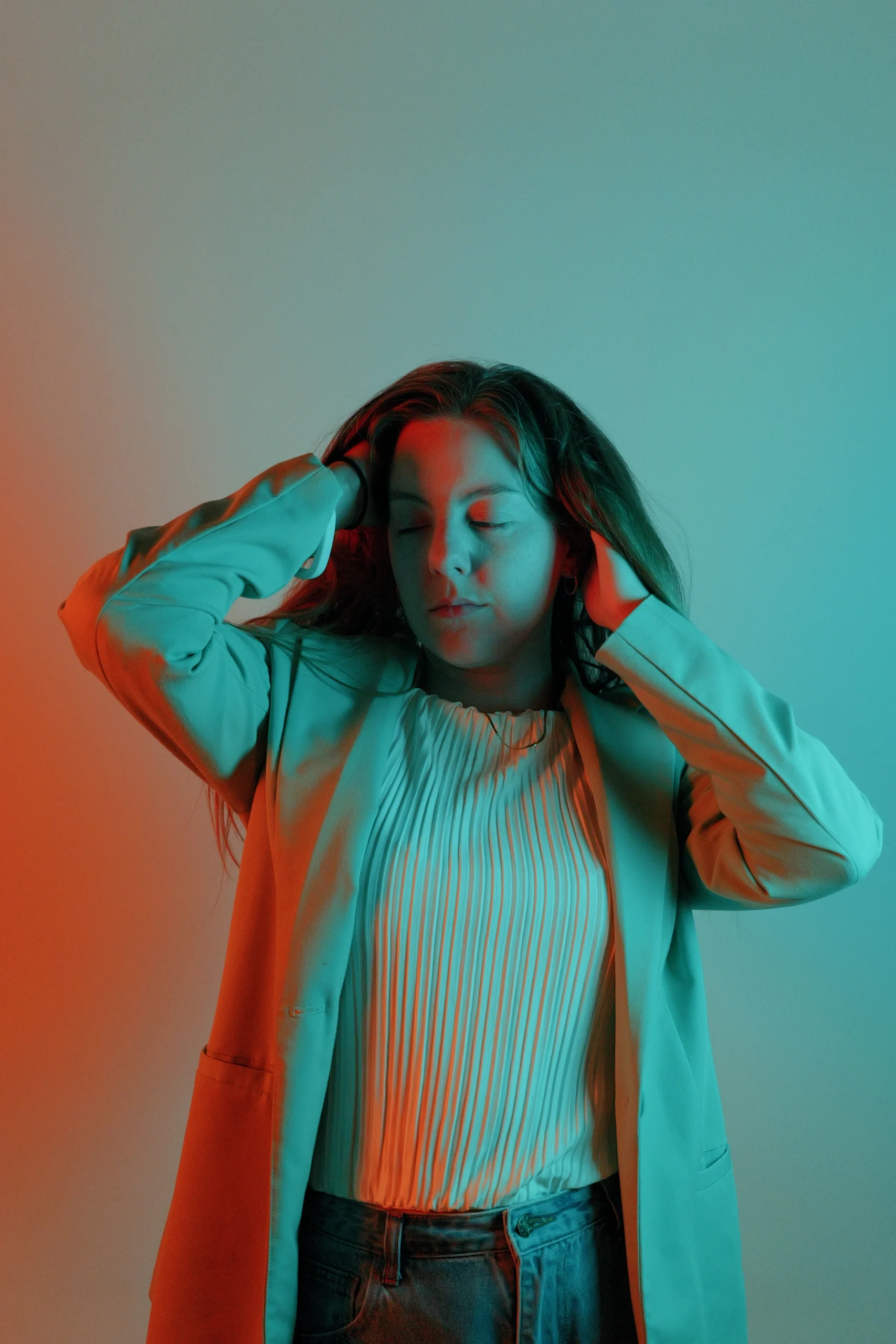Split gel studio portrait in teal blazer and white pleated top, eyes closed with hands in hair