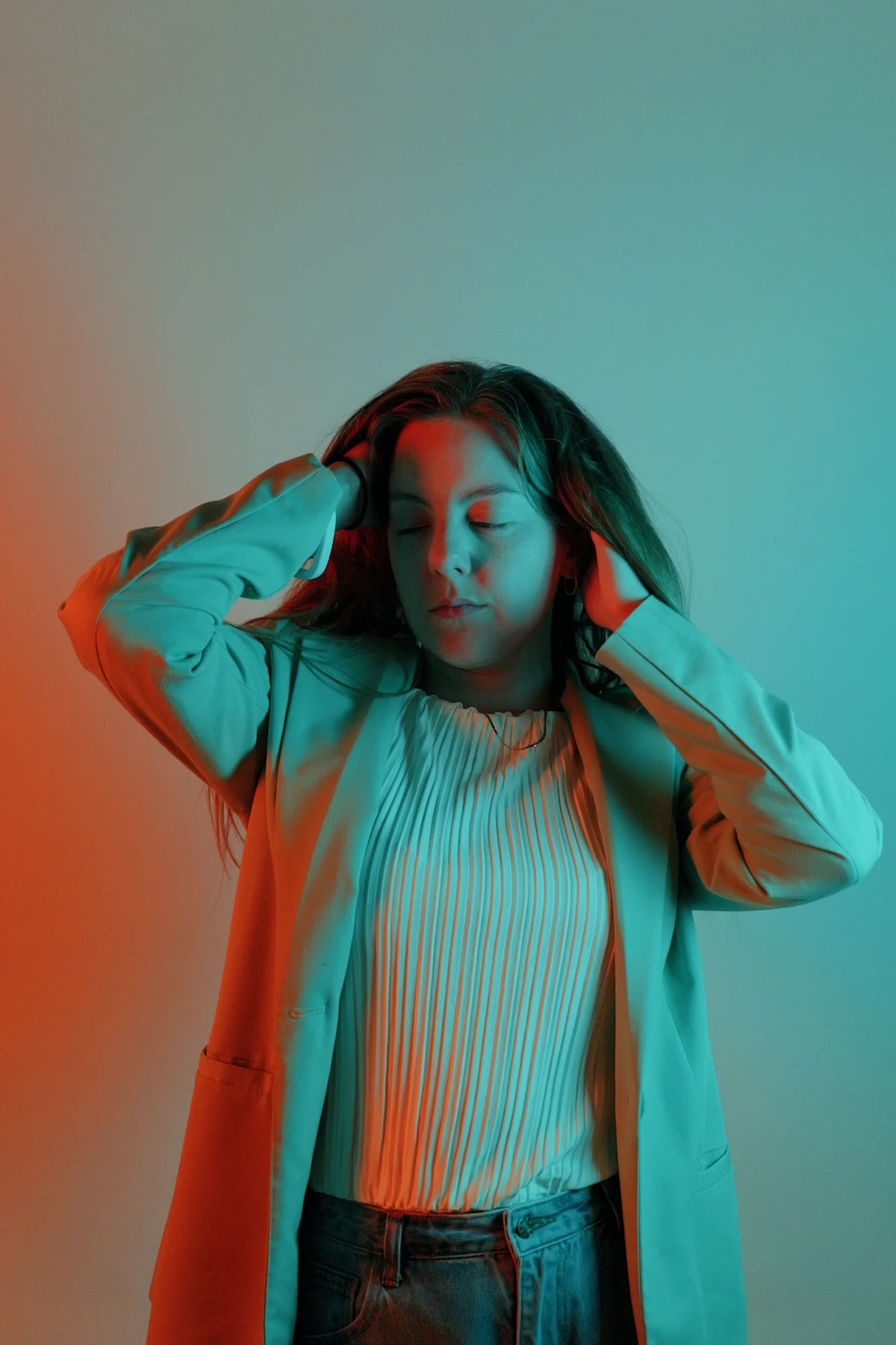 Split gel studio portrait in teal blazer and white pleated top, eyes closed with hands in hair