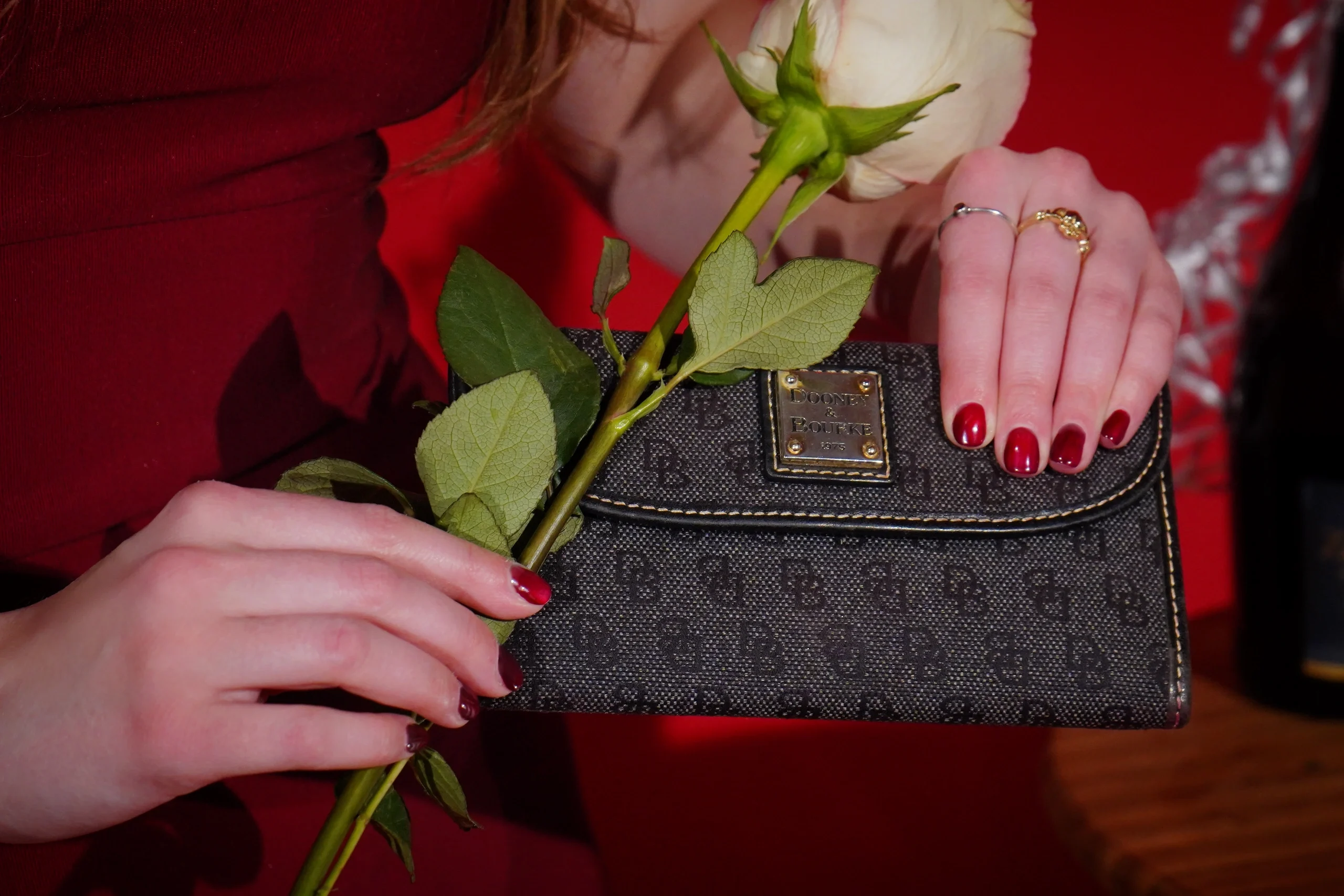 Detail shot of hands holding designer clutch and white roses, red nail polish and gold rings