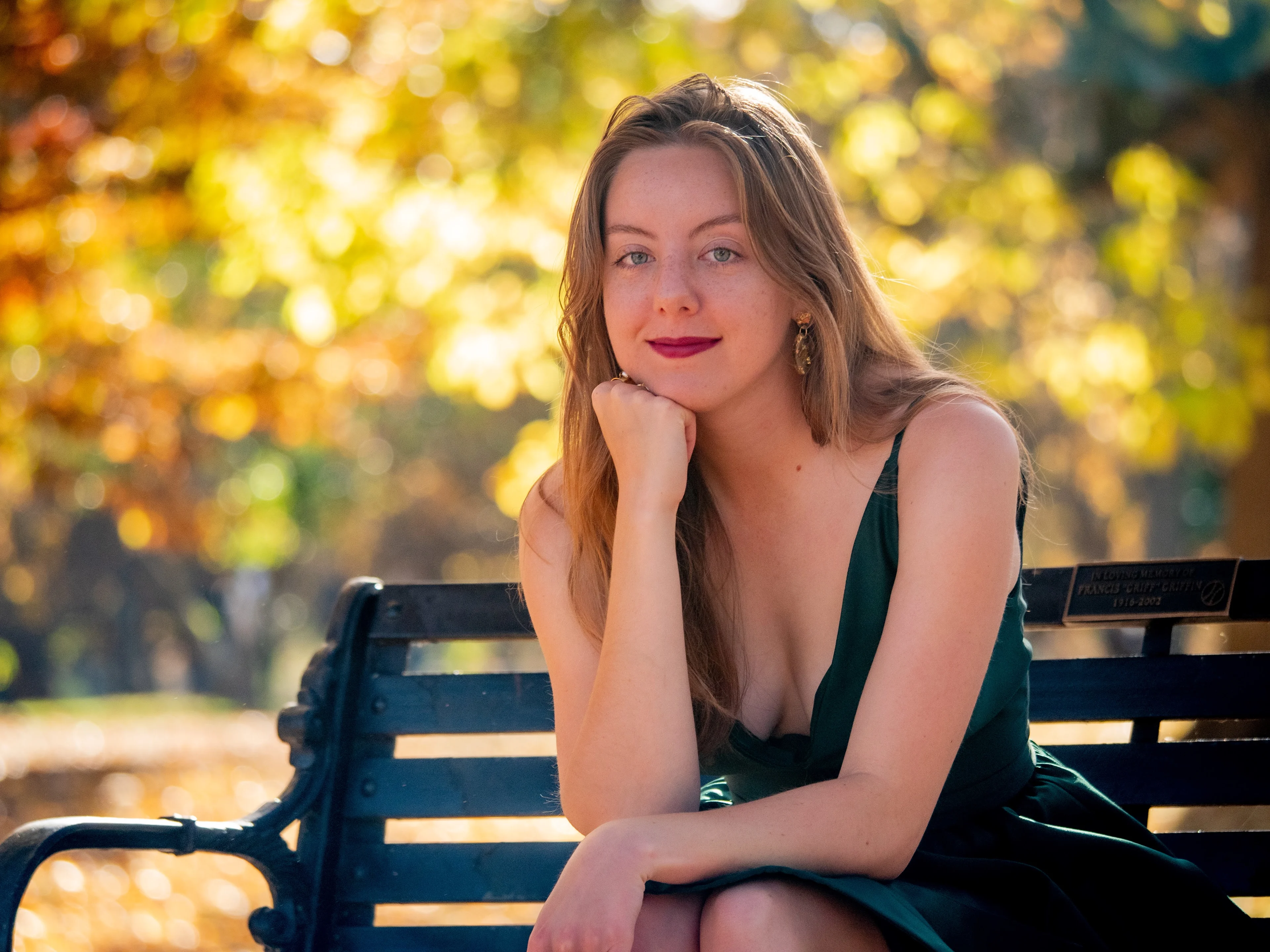 Model on park bench in green dress, hand under chin, warm autumn bokeh background with natural light