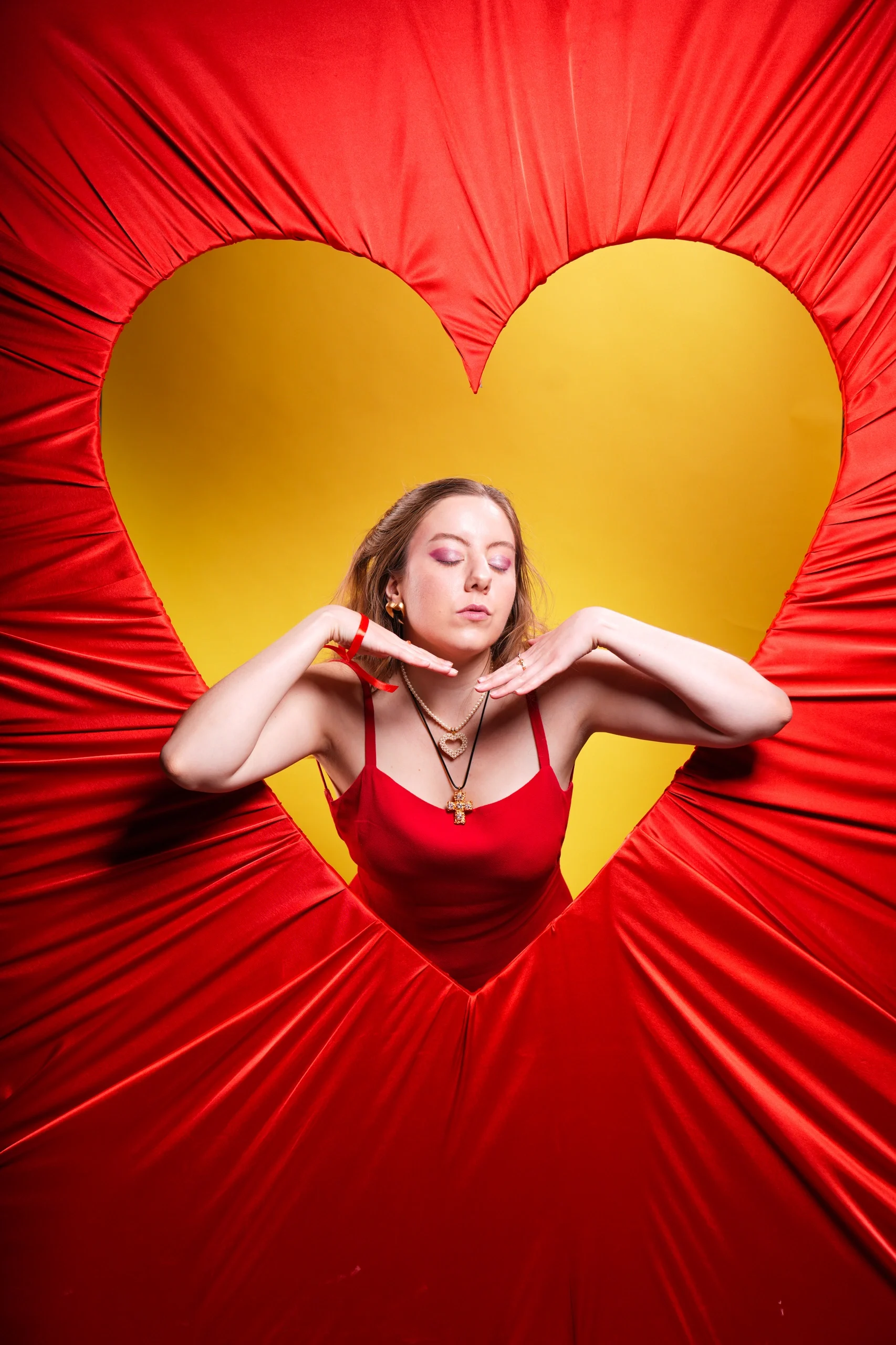 Model in red bodysuit with hands near collarbone, framed by golden heart against red draped fabric