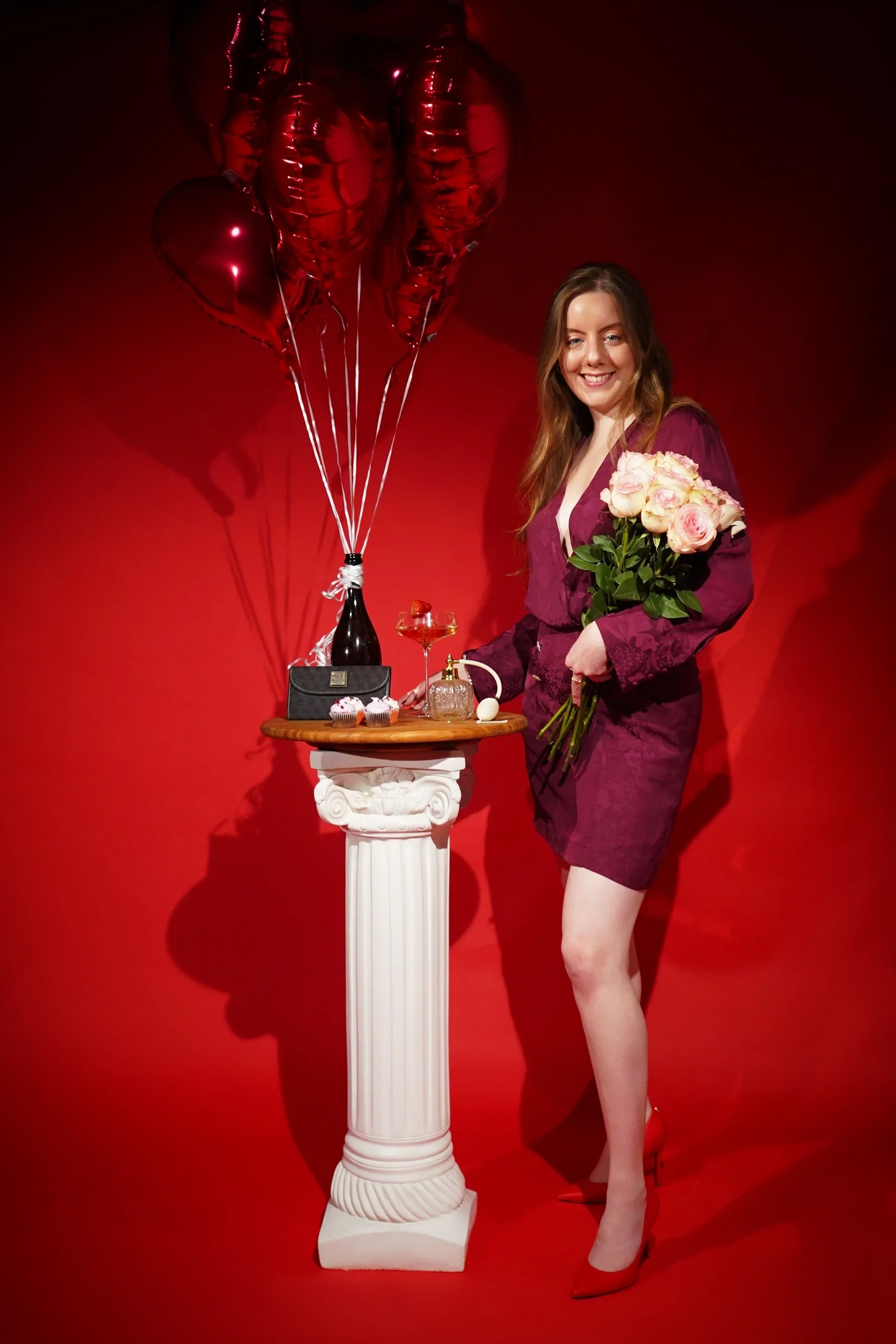 Full-body studio portrait in burgundy dress, holding roses beside white pedestal with balloons