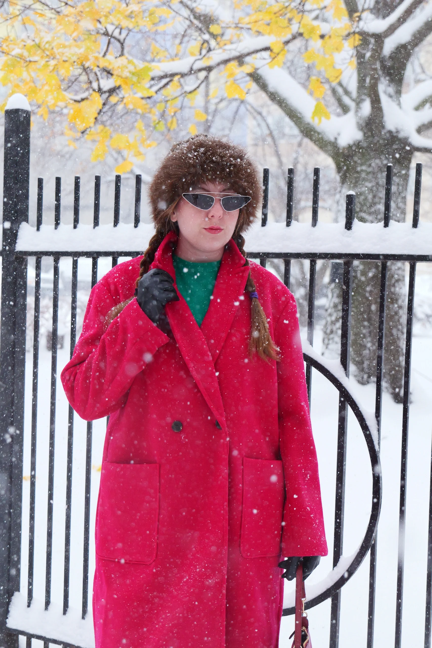 Full-body portrait in red coat, brown fur hat, and sunglasses with iron fence and falling snow