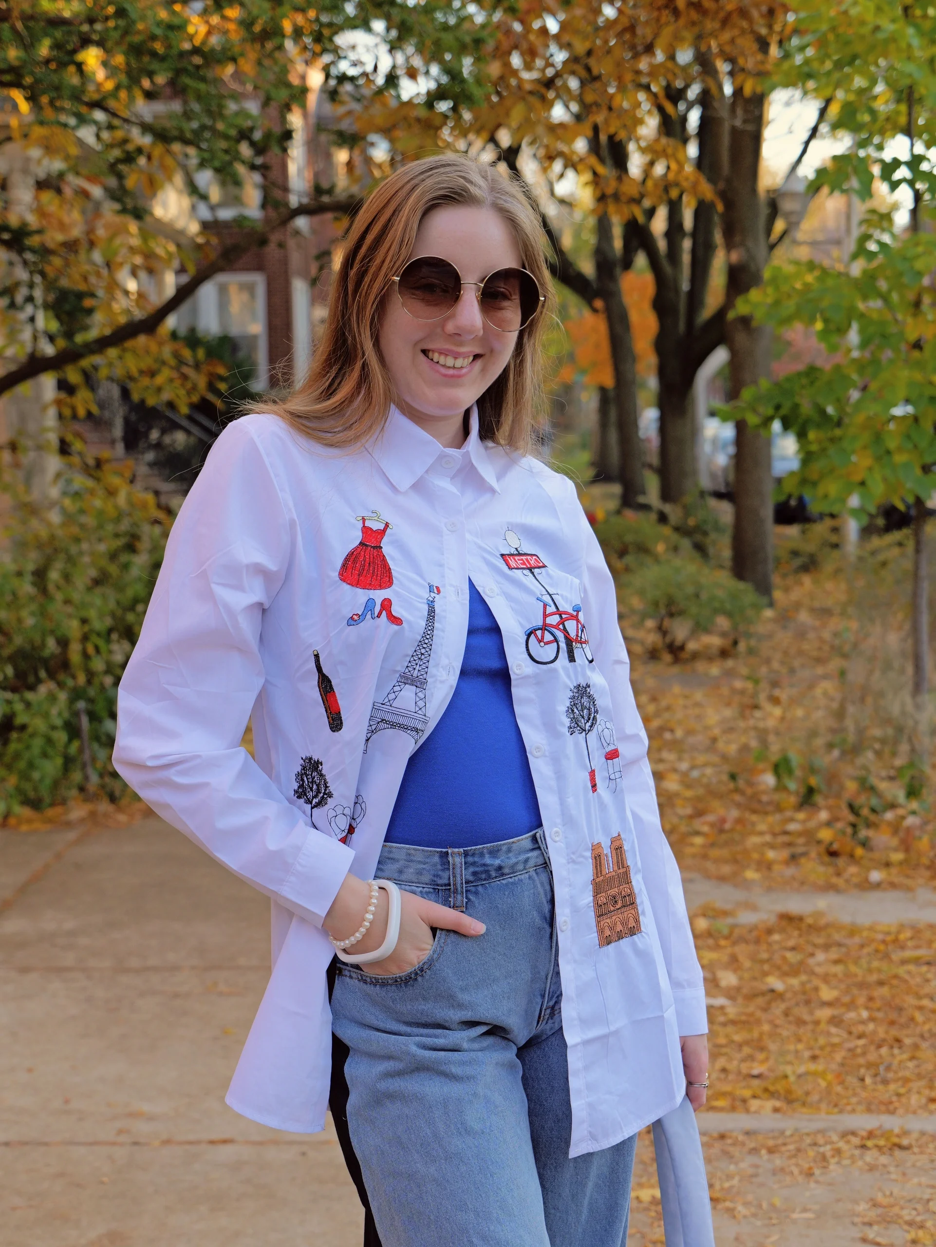 Model in Paris-themed jacket, round sunglasses, blue top, and jeans with autumn park backdrop