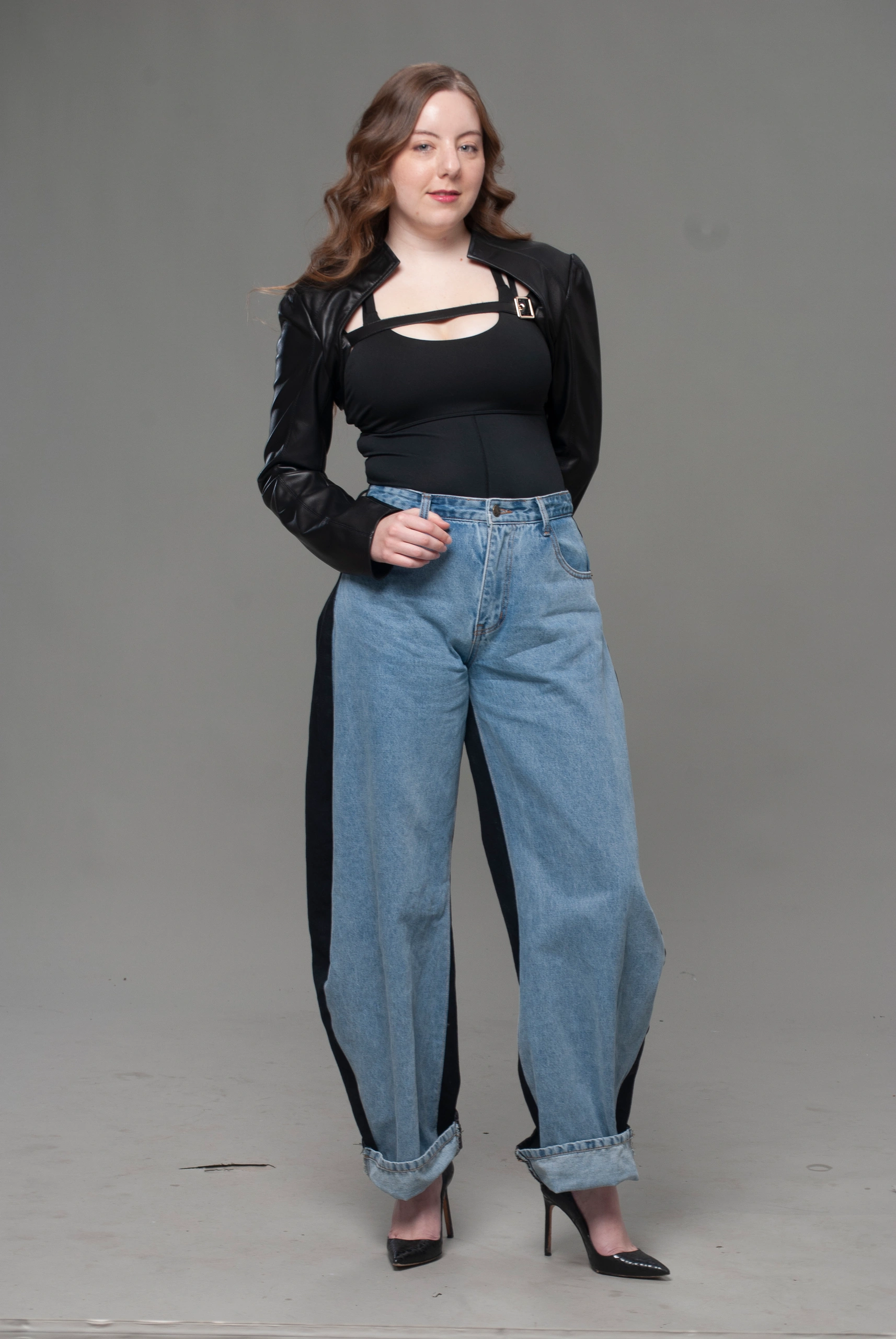 Full-body studio portrait in black coat, cutout top, wide-leg denim jeans, and heels on gray backdrop