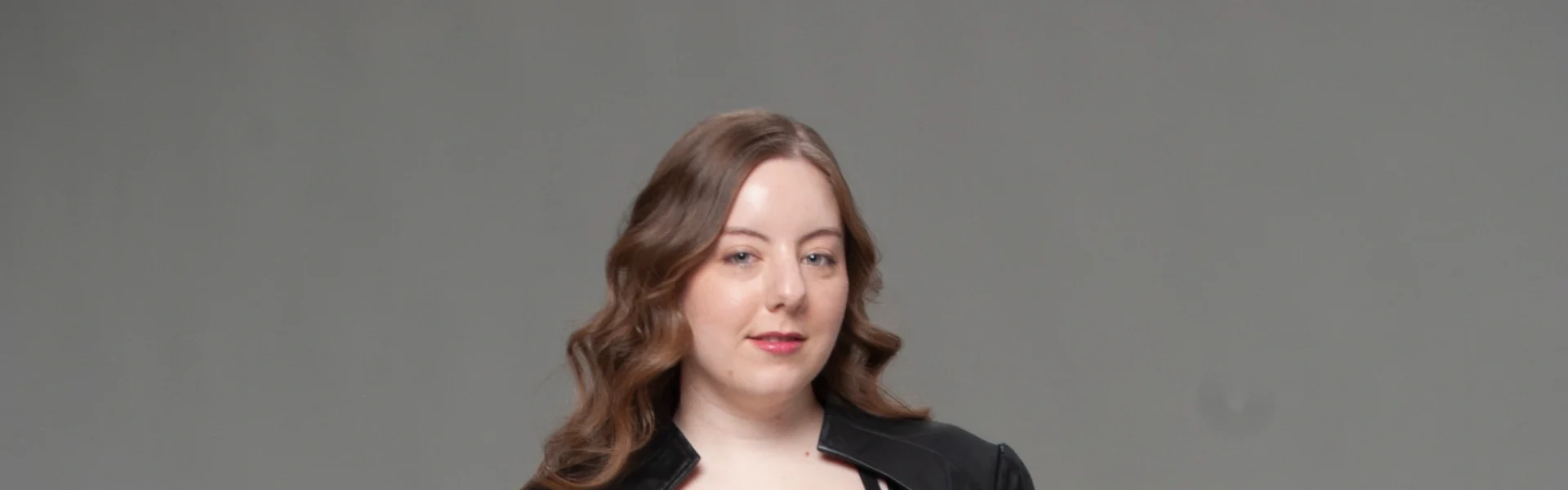 Full-body studio portrait in black coat, cutout top, wide-leg denim jeans, and heels on gray backdrop