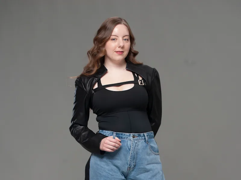 Full-body studio portrait in black coat, cutout top, wide-leg denim jeans, and heels on gray backdrop