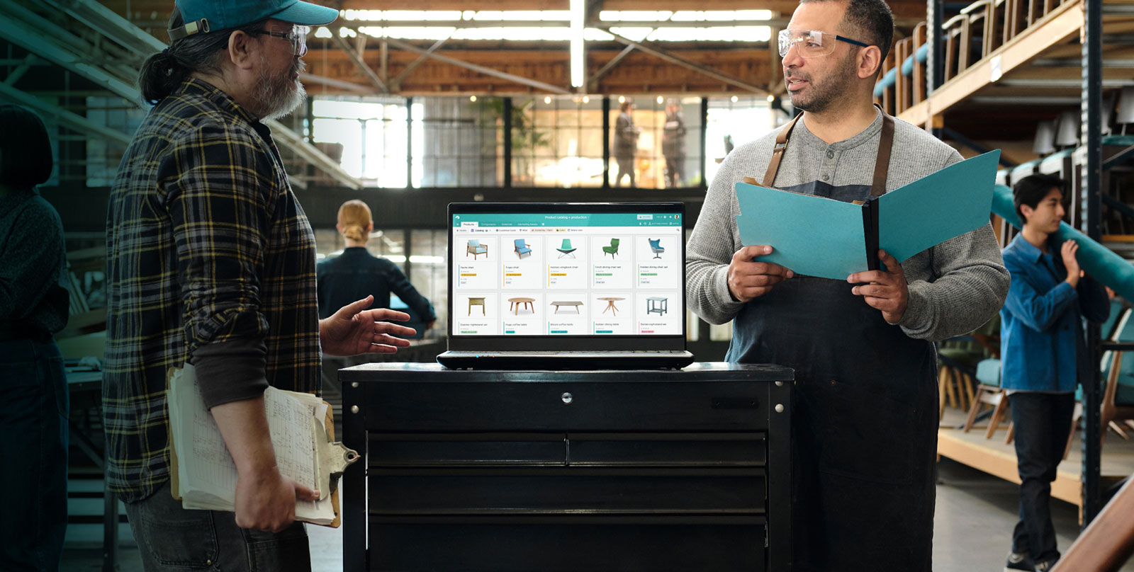 A designer and production staffer talk. A laptop shows chairs tables etc in an Airtable base