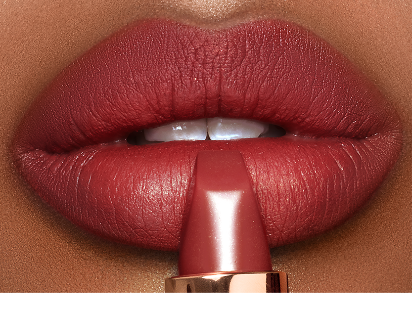 Lips close-up of a deep-tone model wearing berry-rose lipstick with a matte finish while holding the lipstick in front of her lips.