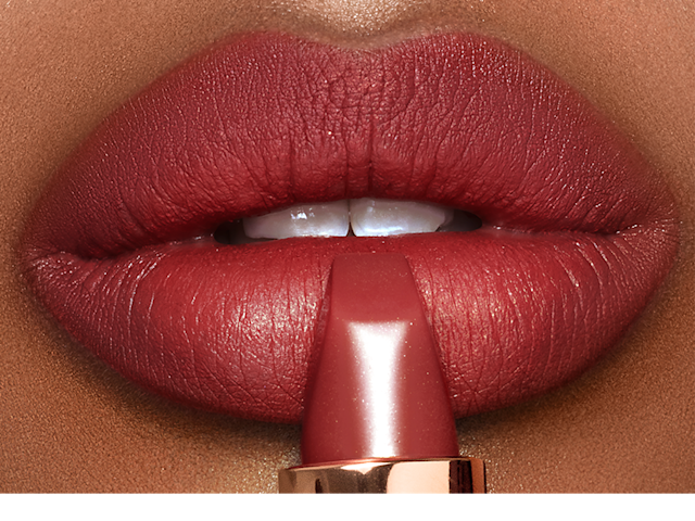 Lips close-up of a deep-tone model wearing berry-rose lipstick with a matte finish while holding the lipstick in front of her lips.