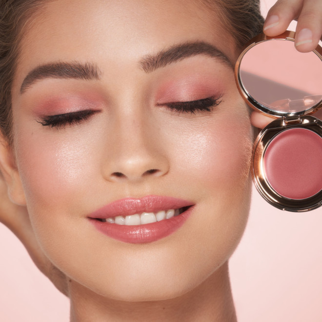 Fair-tone model holding an open, mirrored-lid lip and cheek cream compact in a warm rosebud-pink shade while using it as eyeshadow, lipstick, and blush.