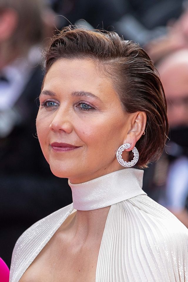 Maggie Gyllenhaal at Cannes Film Festival 2021, wearing a white gown with a low-cut neck with shimmery silver earrings, and soft, glowy nude makeup done using Charlotte Tilbury products.
