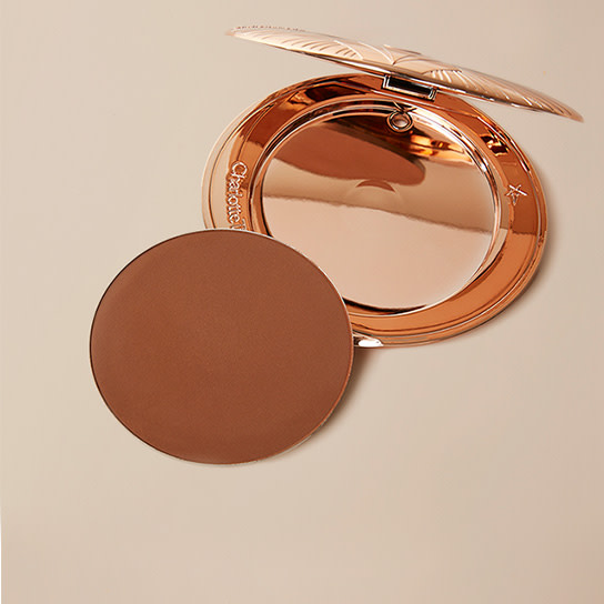 An empty bronzer compact with a mirrored lid with a dark brown-coloured bronzer refill placed next to it.