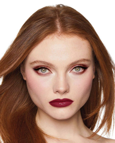 A fair-tone model with grey eyes wearing reddish-plum eye makeup, glowy face base, and a vampy-red lipstick with a satin-finish. 