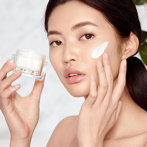 Fair-tone brunette model with glowy, flawless, glass skin, holding an open jar of pearly-white face cream and applying the cream on her face. 