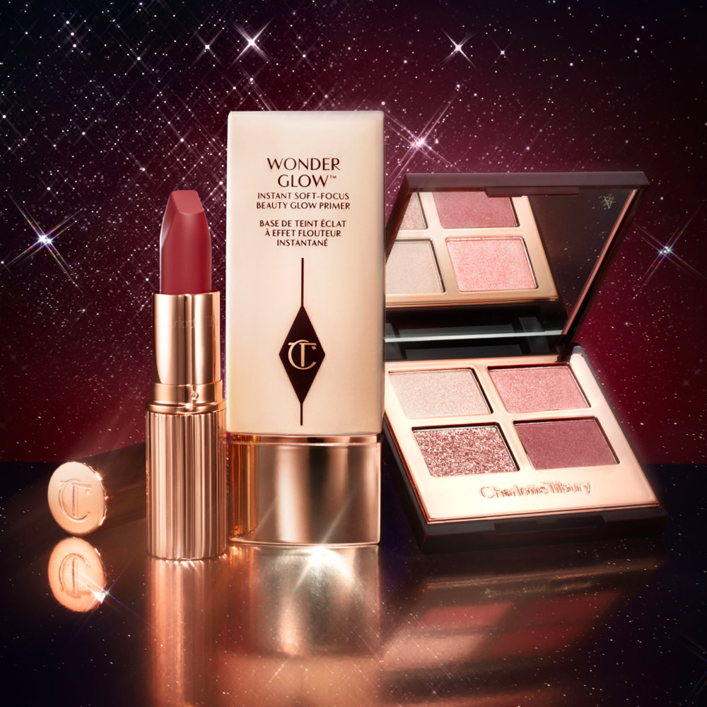 Ruby red matte lipstick in a gold-coloured tube, glowy primer in a clear bottle with a gold-coloured lid, and quad eyeshadow palette with a mirrored-lid in shades of pink and brown.
