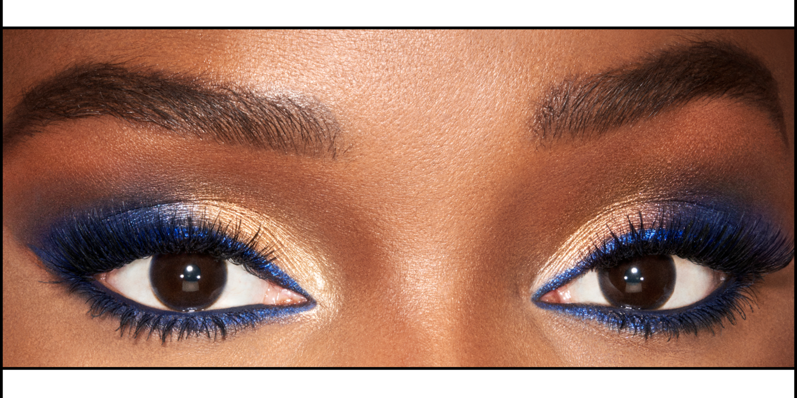 Eyes close-up of a deep-tone model with brown eyes wearing shimmery and metallic royal blue and gold eyeshadow with blue eyeliner on her upper lid and lower waterline.