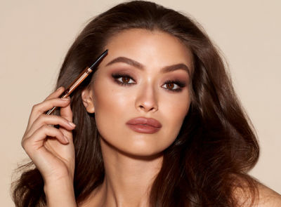 Medium-tone brunette model with brown eyes wearing muted red lipstick with smokey brown eye makeup with lined, filled, and shaped eyebrows and holding an eyebrow pencil in one hand.