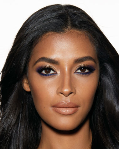 A deep-tone brunette model with brown eyes with shimmery gold and royal blue eyeshadow with matte and metallic eyeliner in royal blue on the upper eyelid and lower waterline. 