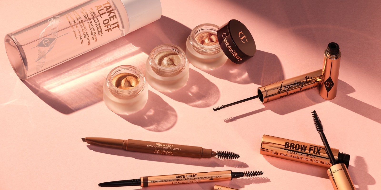 Trio of cream eyeshadows in shimmery shades of gold and pink in open pots with dark brown lids, eyebrow tints, eyebrow gels and brushes, mascara, and makeup remover in a large, clear bottle with a white-coloured cap.