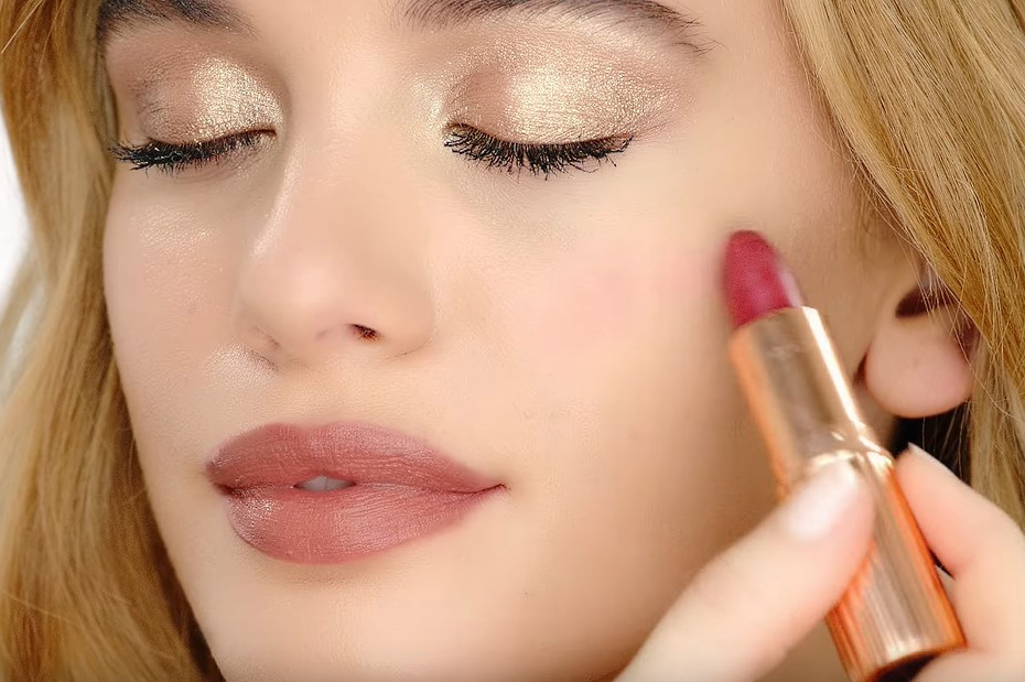 A light tone, blonde model using a warm berry-rose lipstick as a blush while wearing nude brown-pink lipstick and shimmery gold eyeshadow. 