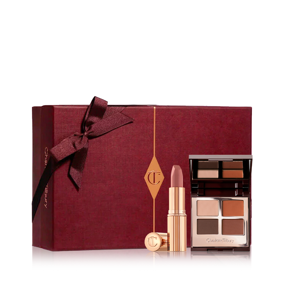 A dark crimson-coloured gift box with an open lipstick in a nude pink shade with an open mirrored-lid quad eyeshadow palette with brown, red, and beige eyeshadows. 