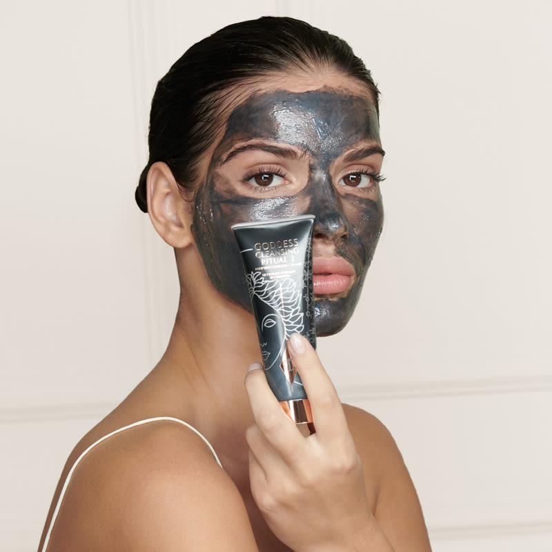A light-tone model wearing a wash-off charcoal mask while holding the jet-black charcoal cleanser. 