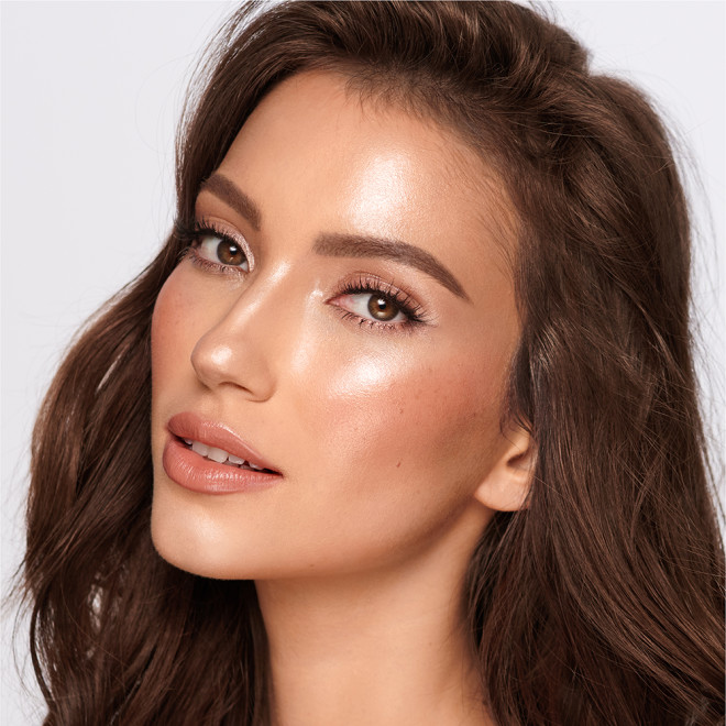 Medium-tone brunette model wearing glossy peach lipstick with shimmery gold and fawn eye makeup, and glowy coral-peach blush.