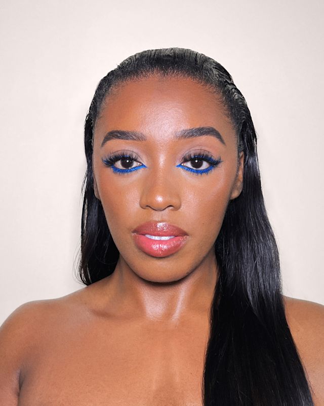 Close-up of a deep-tone model with brown eyes wearing soft pinkish-peach glossy lipstick with brownish-pink blush, and brown and beige eye makeup with blue eyeliner on her lower waterline.