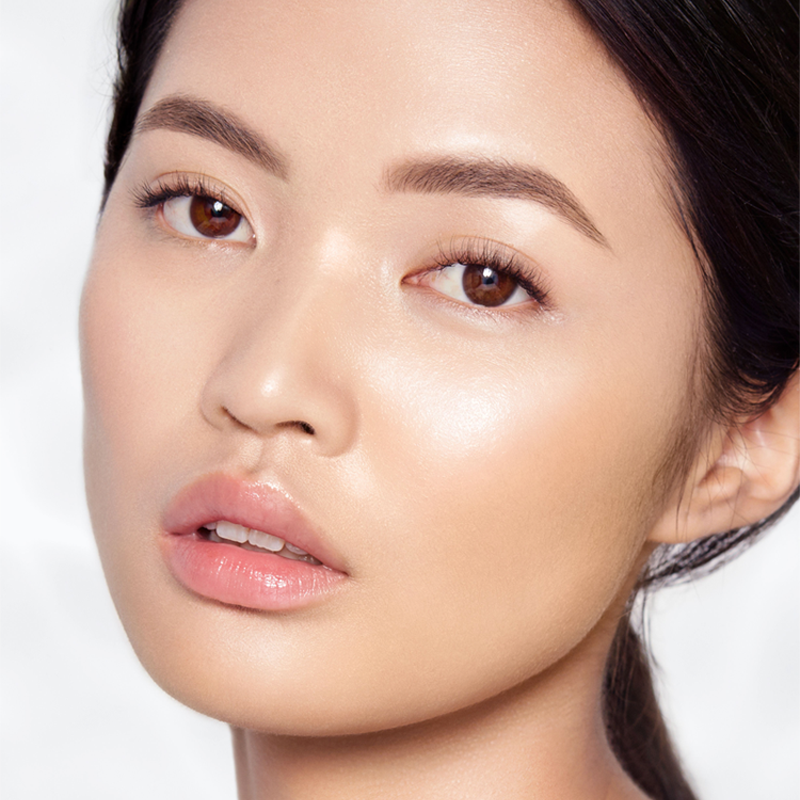 Fair-tone brunette model with flawless, glowy skin, wearing dark brown eyeliner with nude pink lip gloss. 