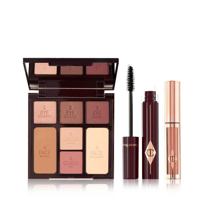 An open, mirrored-lid face palette with nude eyeshadows, bronzer, blushes, and highlighter, an open mascara in dark crimson packaging with its applicator next to it, and nude pink liquid lipstick in rose-gold packaging. 