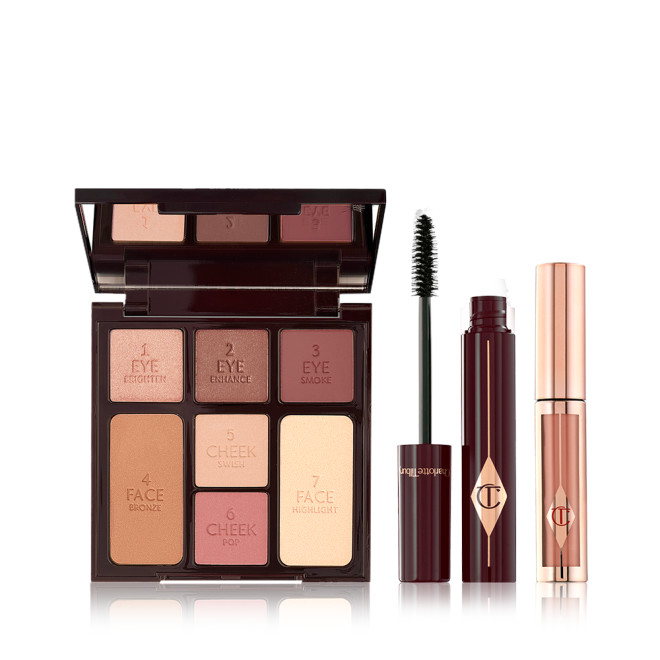 An open, mirrored-lid face palette with nude eyeshadows, bronzer, blushes, and highlighter, an open mascara in dark crimson packaging with its applicator next to it, and nude pink liquid lipstick in rose-gold packaging. 