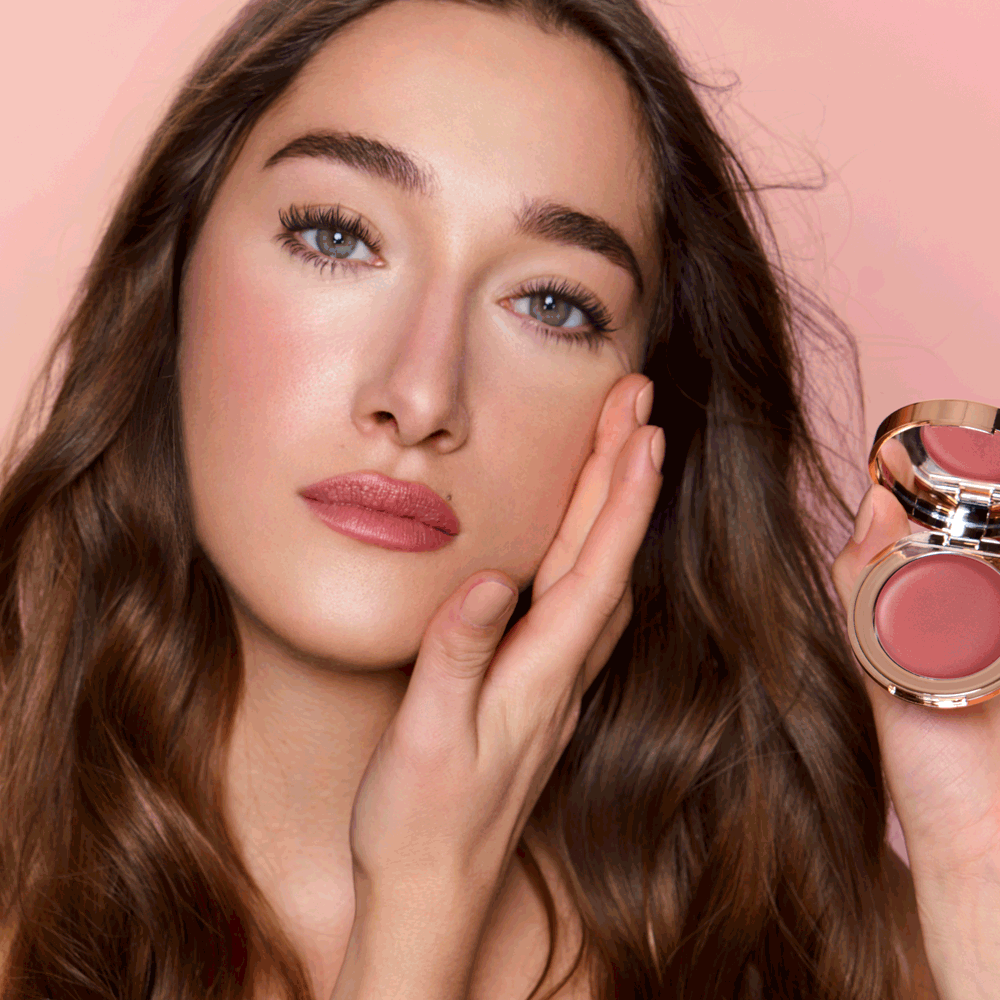 Close-up of a fair-tone model with blue eyes wearing a lip and cheek cream blush in a glowy deep berry-pink colour on her lips and cheeks while holding the lip and cheek compact beside her face.
