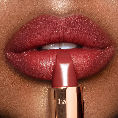 Lips close-up of a deep-tone model wearing dark berry-pink lipstick while holding the lipstick in front of her lips.