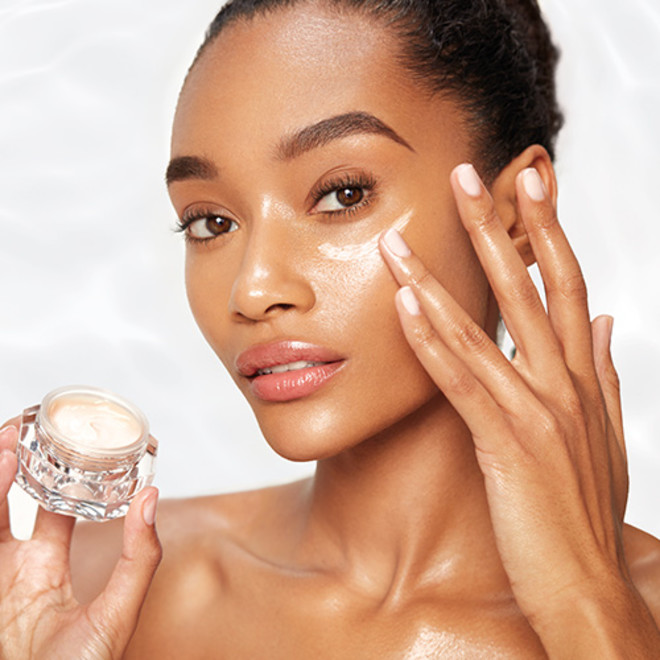 A deep-tone brunette model with glowy, flawless skin applying a fawn-coloured, nourishing eye cream and holding the petite eye cream jar in her other hand.