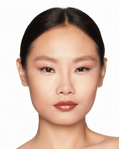 Fair-tone model with brown eyes wearing a moisturising lipstick balm in a peachy-nude shade with a high-shine finish.