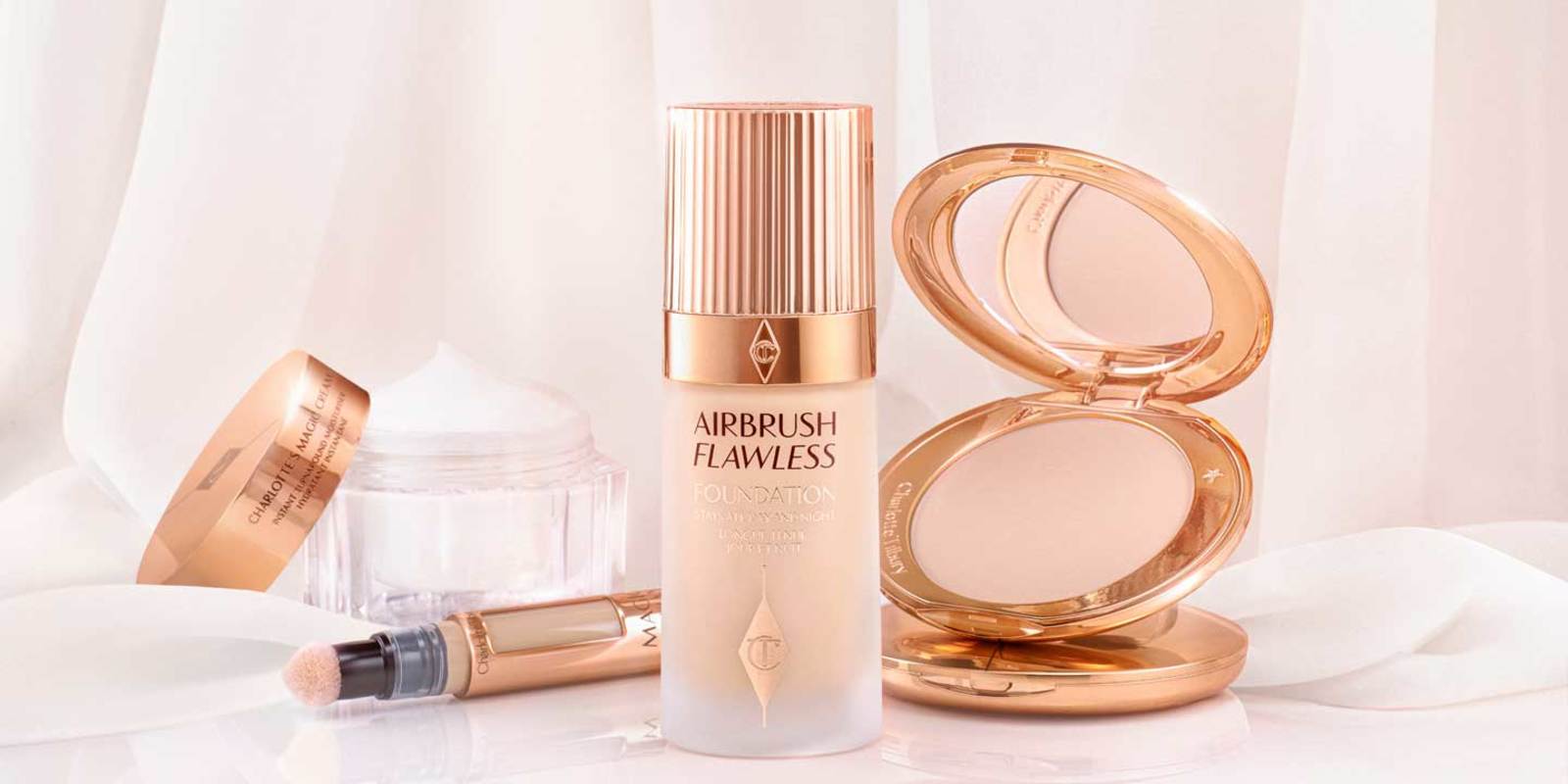 An open face cream in a glass jar with a rose-gold coloured lid, an open concealer, foundation in a frosted glass bottle with a rose gold coloured lid, and an open, mirrored-lid powder compact. 