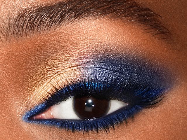 Eye close-up of a deep-tone model with brown eyes wearing shimmery gold and blue eyeshadow with matte and metallic blue eyeliner on the upper lid and waterline.