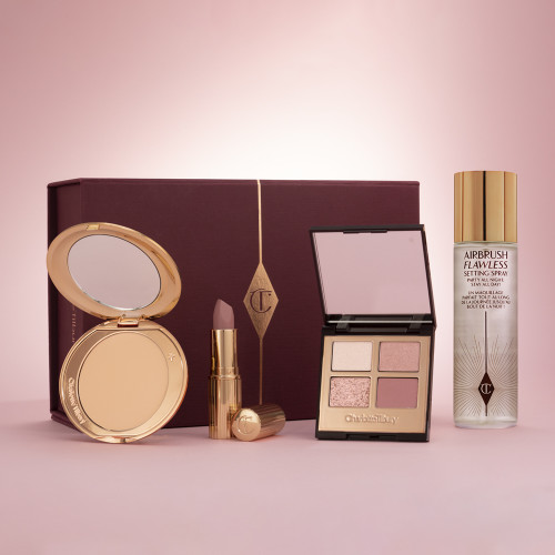 A dark scarlet-coloured gift box with a pressed powder compact with a mirrored-lid, an open lipstick in a mauve shade, a quad eyeshadow palette with pink and gold shades, and a setting spray. 