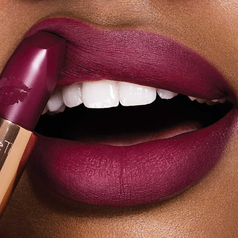 Lips close-up of a deep-tone model applying a deep purple berry matte lipstick.