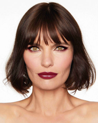 A medium-light-tone model with hazel eyes wearing reddish-plum eye makeup, glowy face base, and a vampy-red lipstick with a satin-finish. 