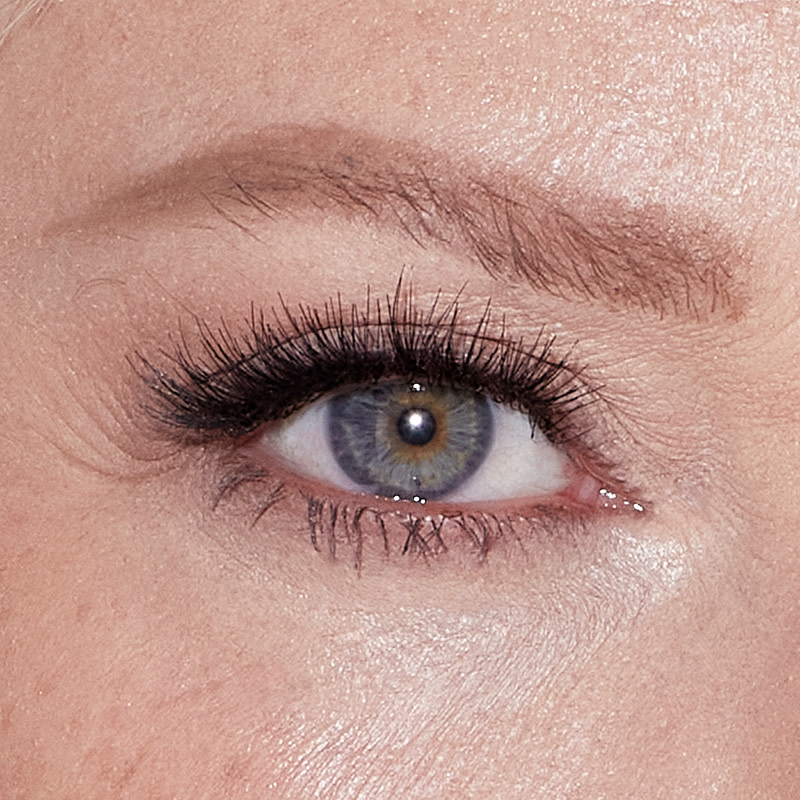 Single-eye close-up of a mature model with grey eyes demonstrating how to fill in eyebrows.