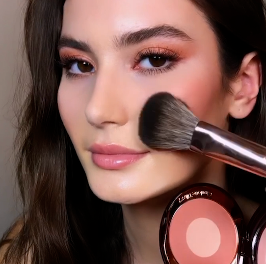 A fair-tone, brunette model wearing nude-pink makeup and getting a glowy, nude-pink blush applied. 