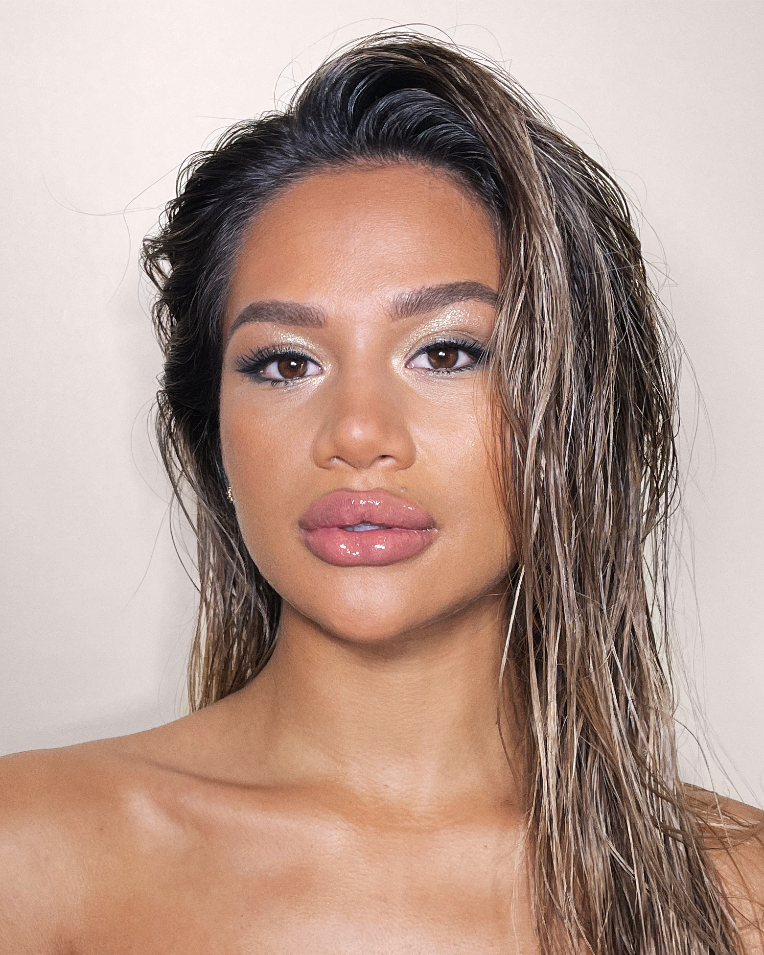 Close-up of a medium-dark-tone model with brown eyes wearing nude pink lipgloss with metallic beige eye makeup, and bronzed skin.