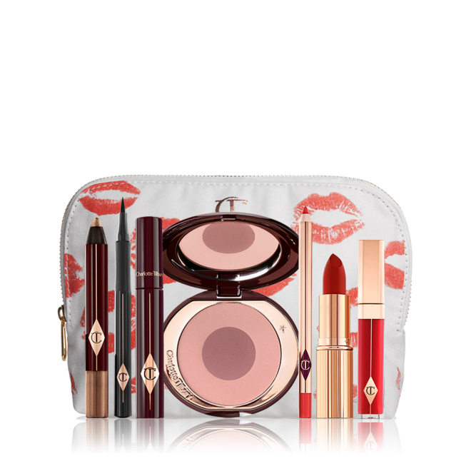 A white makeup pouch with an open two-tone blush in cool-toned pink and brown with a mascara, eyeliner pen, chubby eyeshadow stick in gold, an open lipstick in bold red, lip liner pencil in blood-red, and a lip gloss in bright red. 