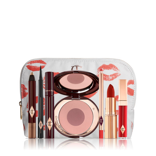 A white makeup pouch with an open two-tone blush in cool-toned pink and brown with a mascara, eyeliner pen, chubby eyeshadow stick in gold, an open lipstick in bold red, lip liner pencil in blood-red, and a lip gloss in bright red. 