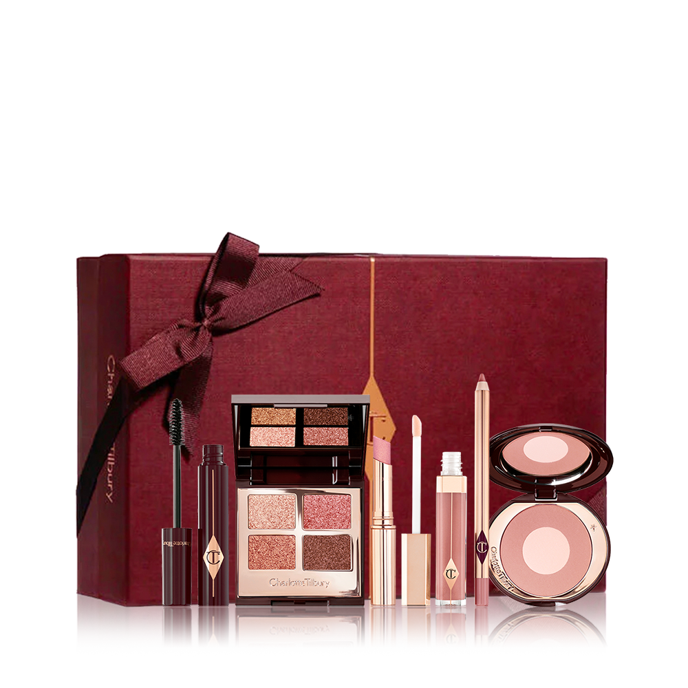 An open mascara tube with the applicator next to it with an open, mirrored-lid quad eyeshadow palette in shimmery neutral shades, a shimmery nude pink lipstick, a nude pink lip gloss and lip liner pencil, and a two-tone powder blush compact in a nude pink shade, with all products in front of a maroon-colour gift box. 